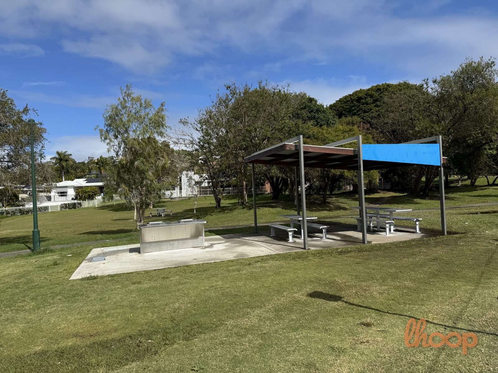 A stainless barbecue and a picnic shelter with a blue roof on the open parkland lawn, with houses and trees beyond.