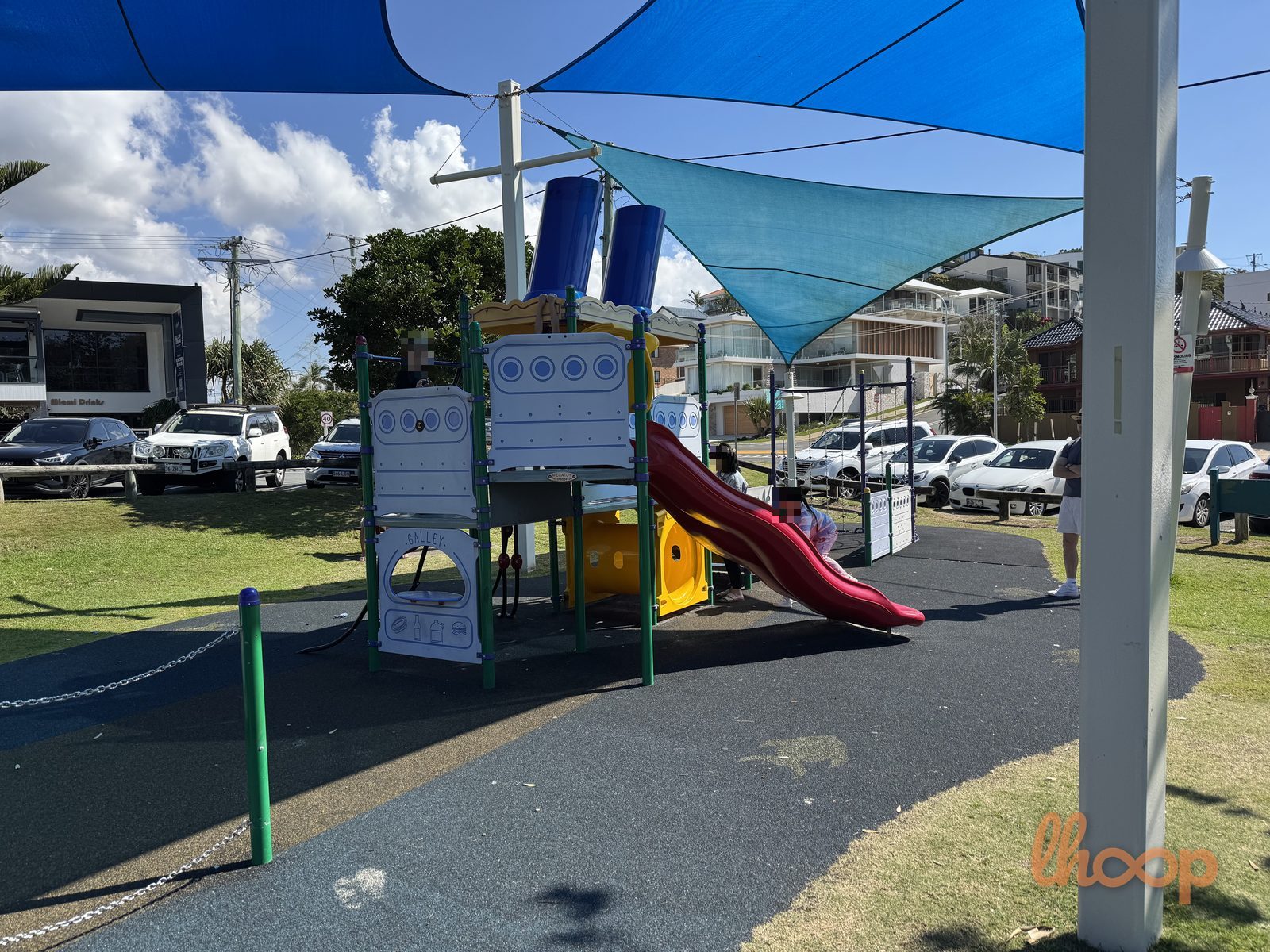 The smaller play tower with a red wave slide under blue shade sails, with parked cars and apartments across the road.