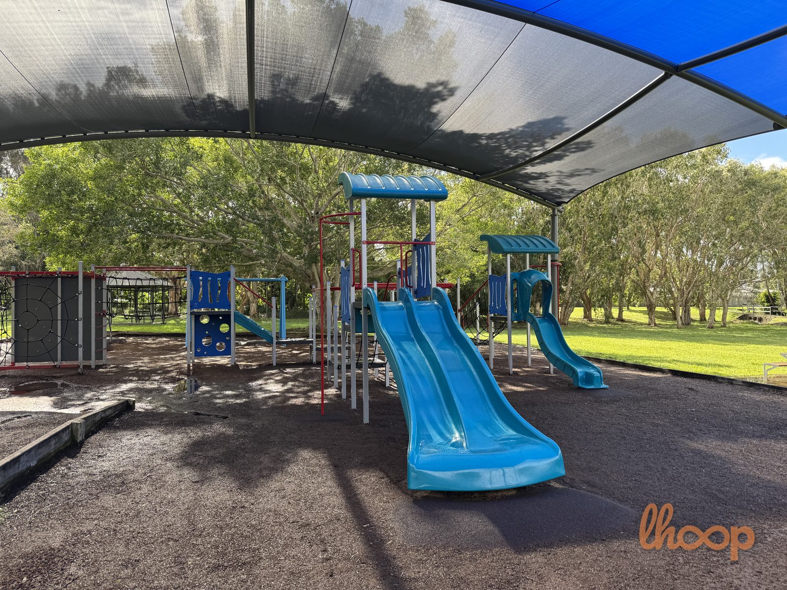 Blue slides and a multi-level play structure with climbing panels and monkey bars under the shade sail.