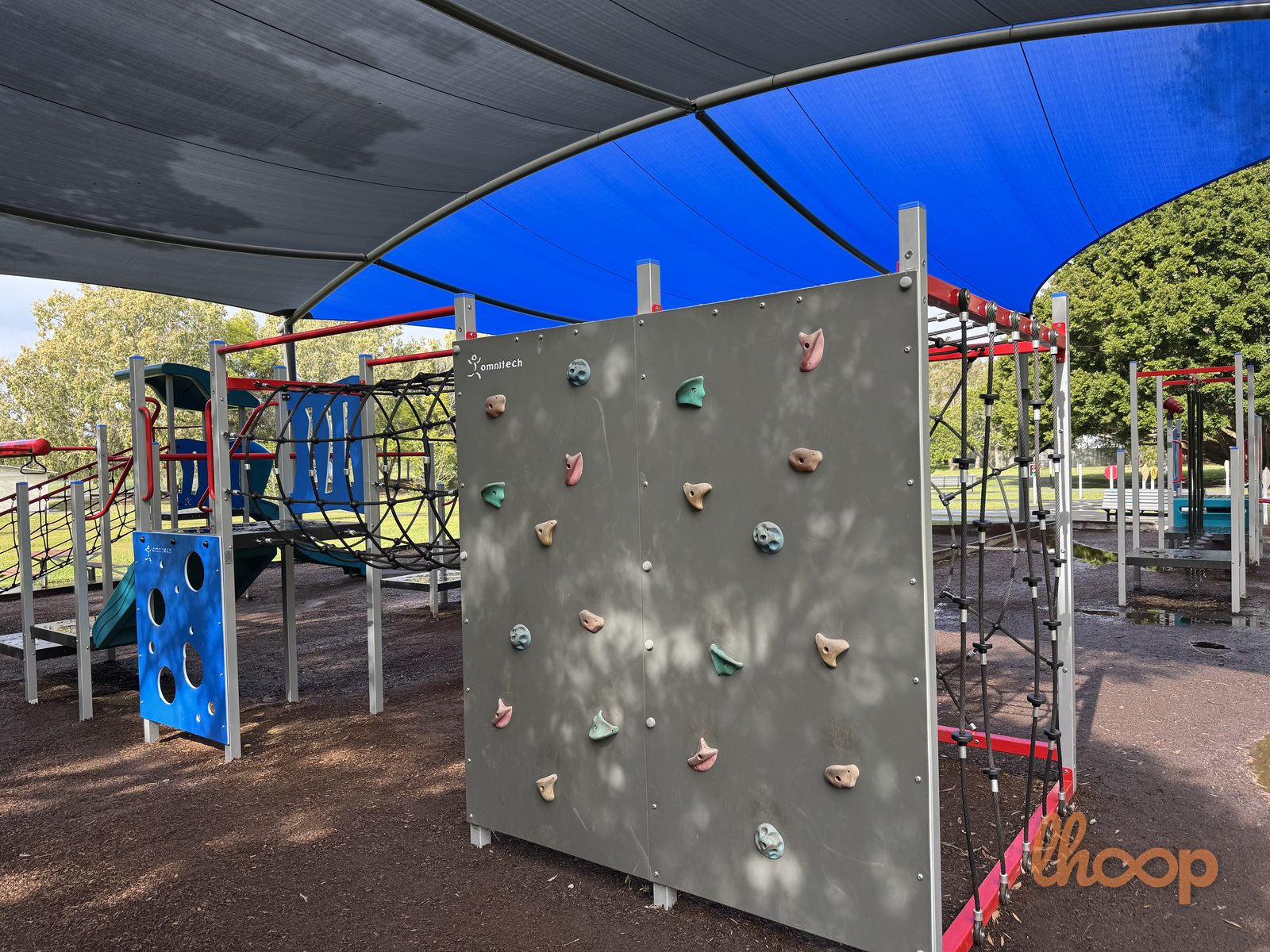 A climbing wall with coloured holds and monkey bars, part of the shaded play structure.