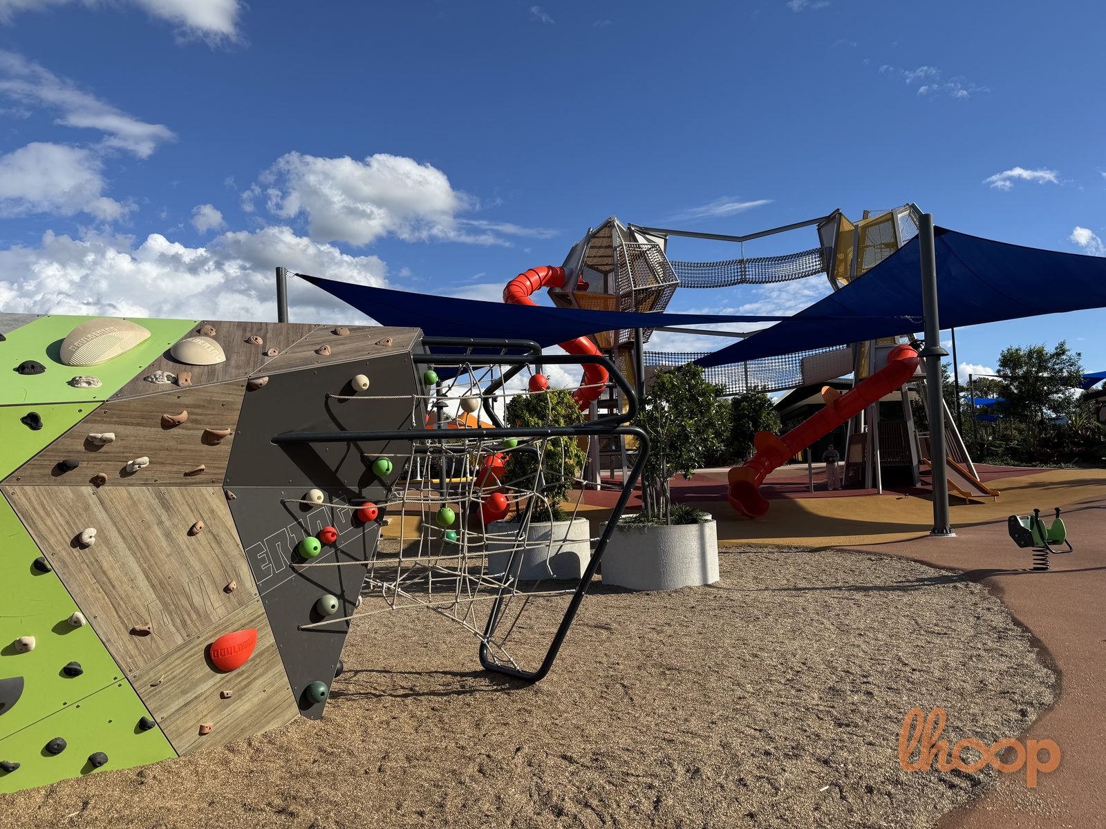 The main climbing structure with a rock wall and rope nets over soft sand, under blue shade sails.