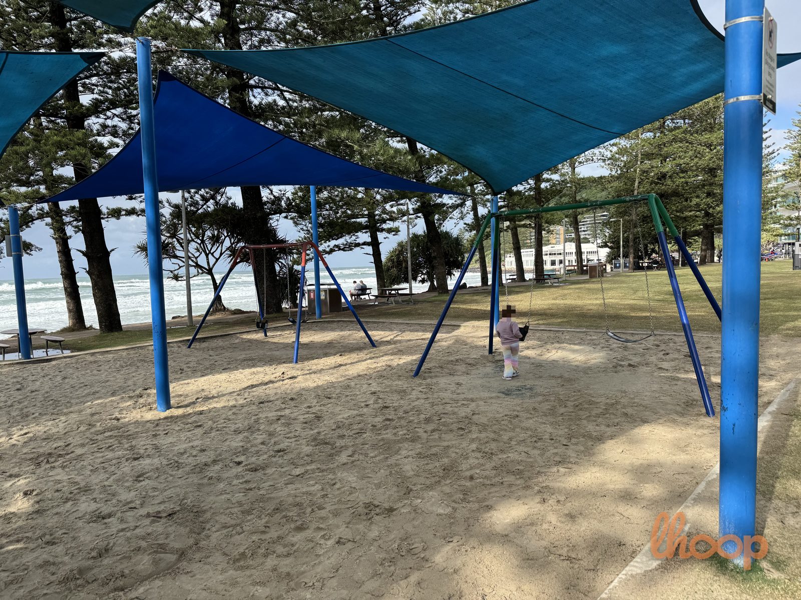 A swing set on the sand under blue shade sails, with the surf and a picnic table beyond.