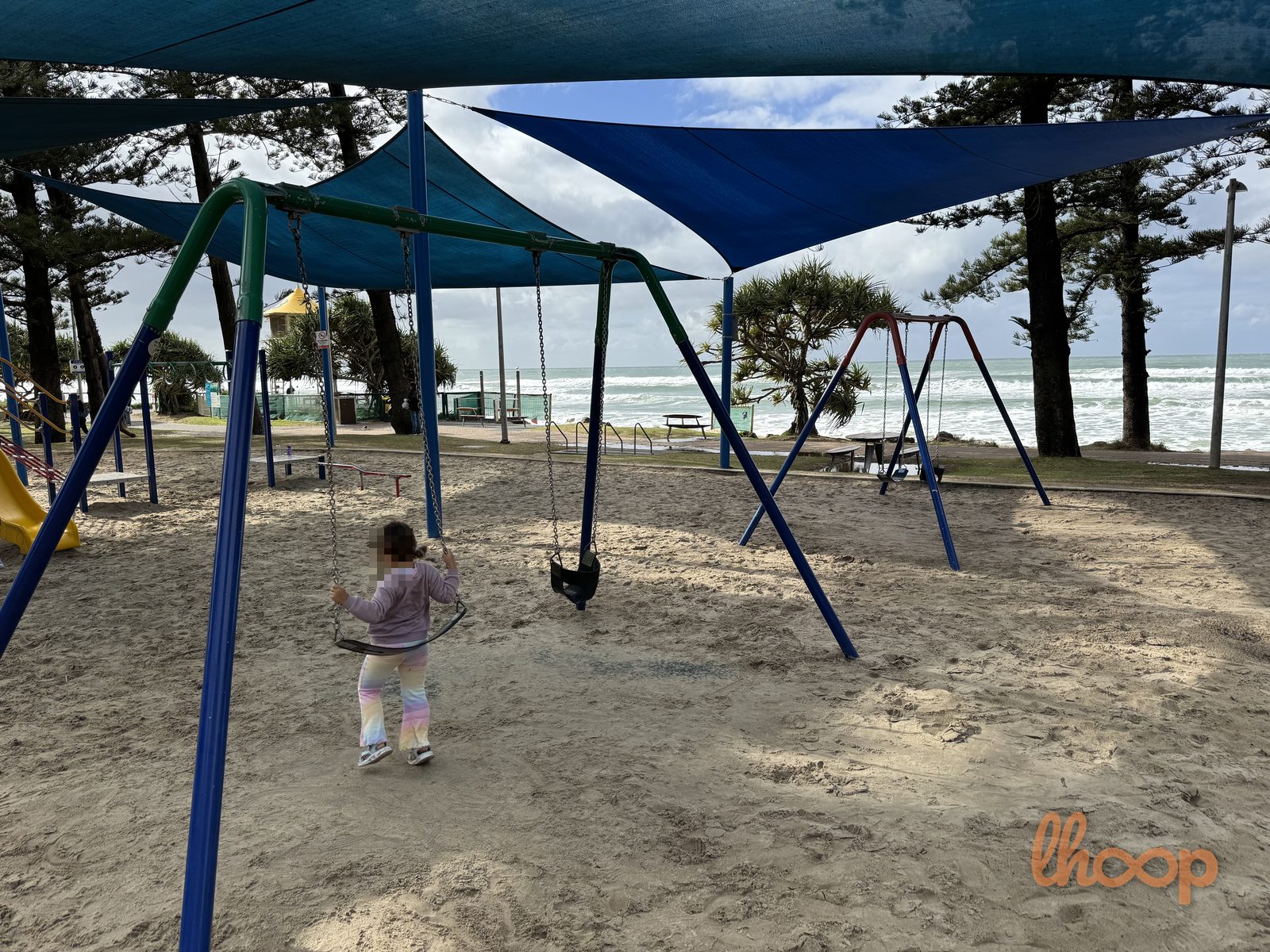 Swings under the shade sails with the ocean and Norfolk pines behind, and a slide unit to the left.