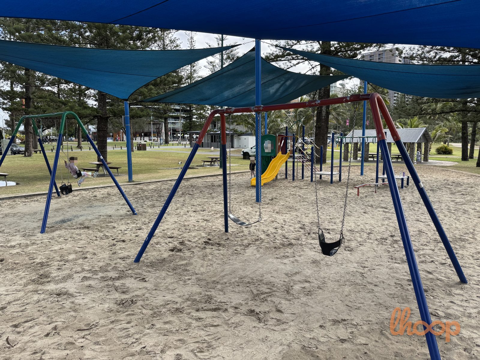 The blue swing set and a yellow slide tower on the sand, framed by Norfolk pines and the sea.