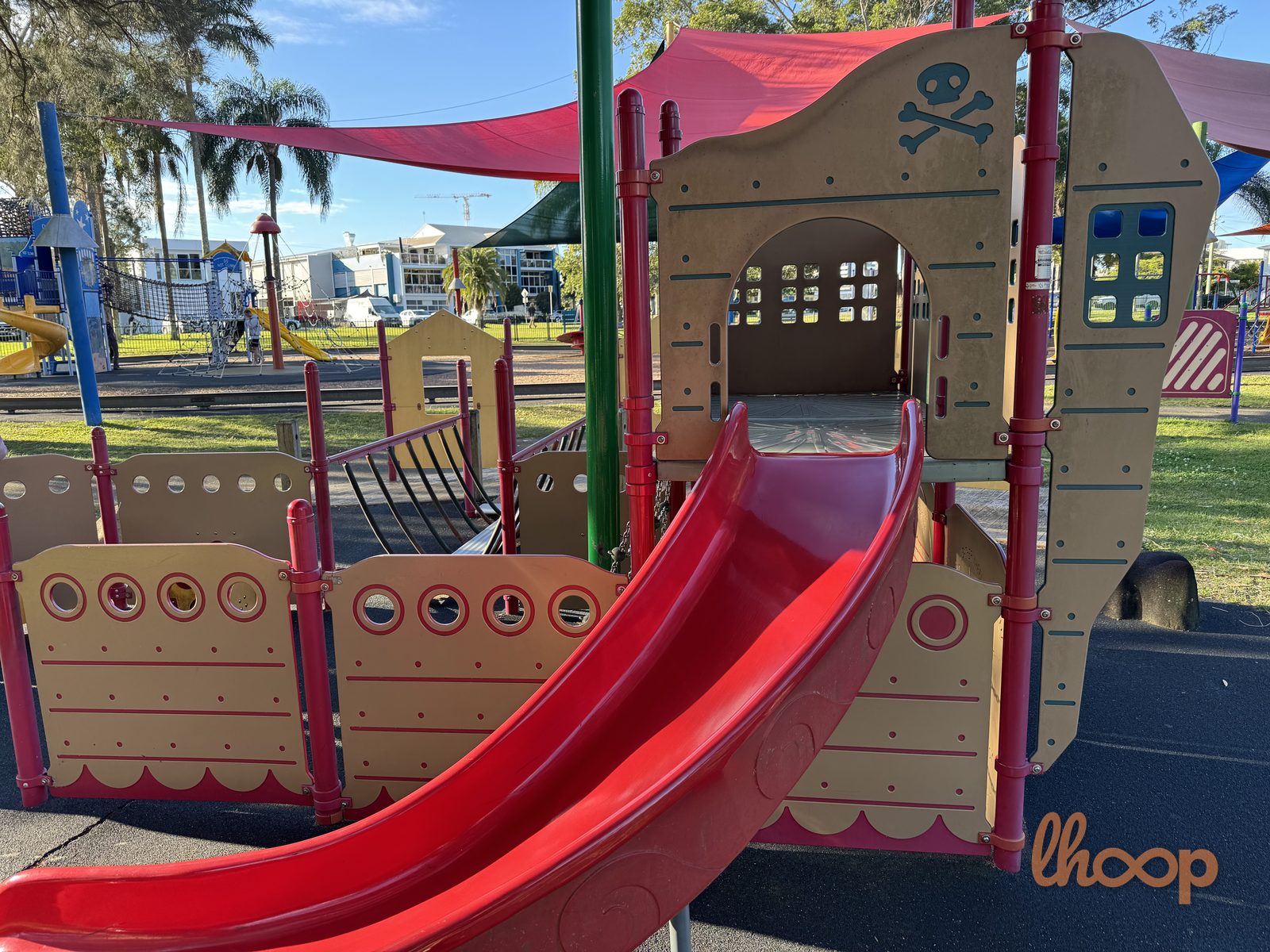 The tan and red pirate ship with a wide red slide, porthole panels and a skull-and-crossbones cut-out, under a red shade sail.