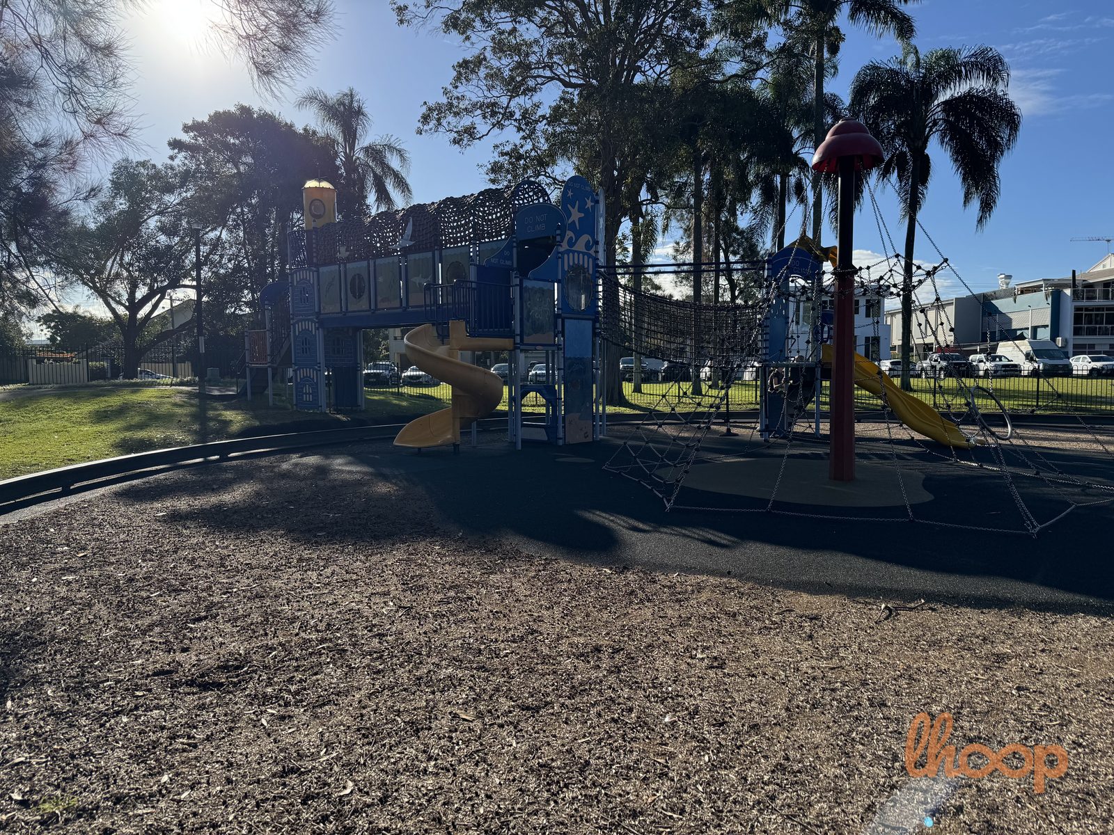 The main play tower seen across the mulch, with slides, a rope net bridge and palms behind the perimeter fence.
