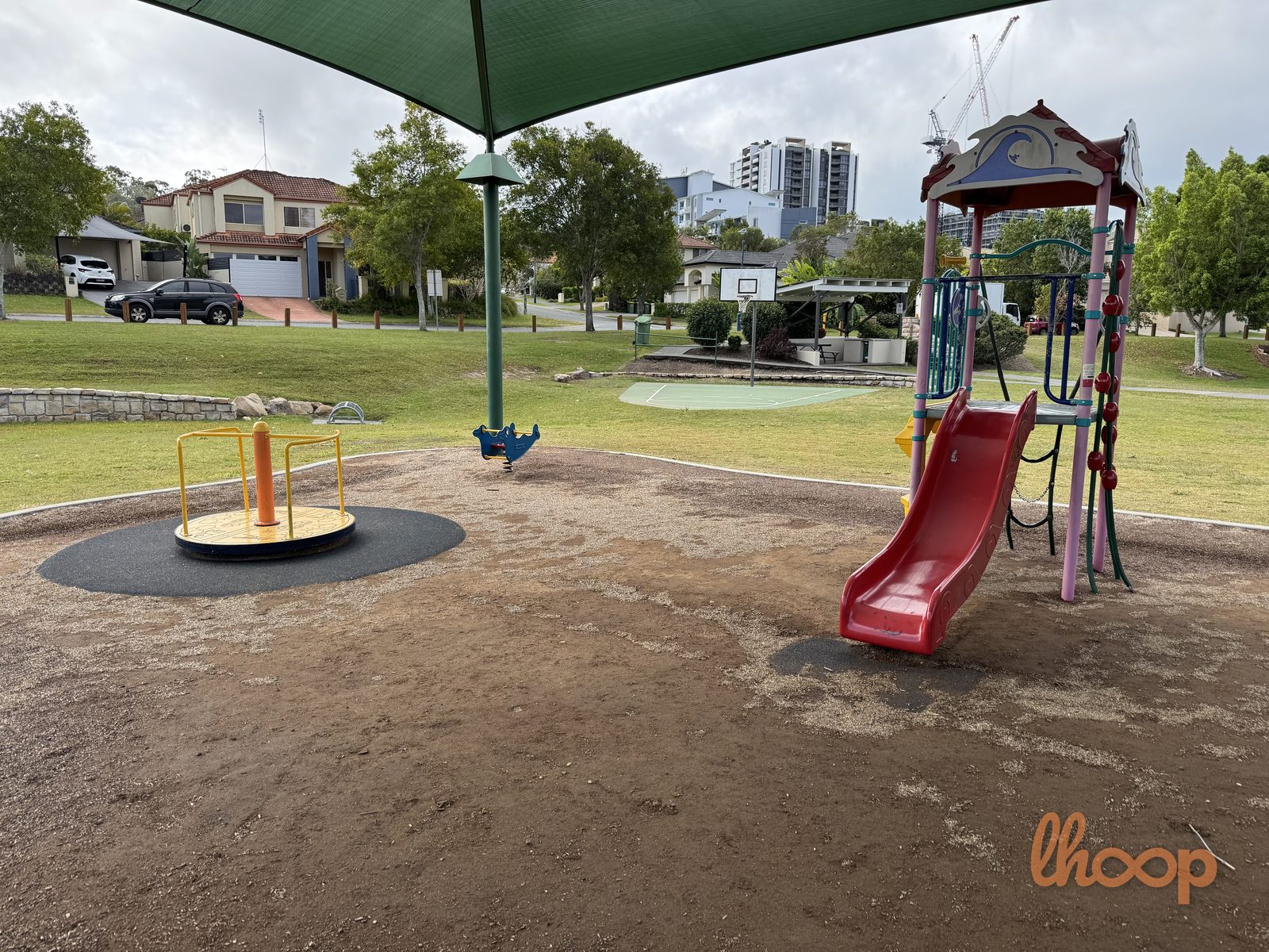 The playground under a green shade sail with an ornate red slide tower, a roundabout and a spring rocker, and a basketball half-court behind.