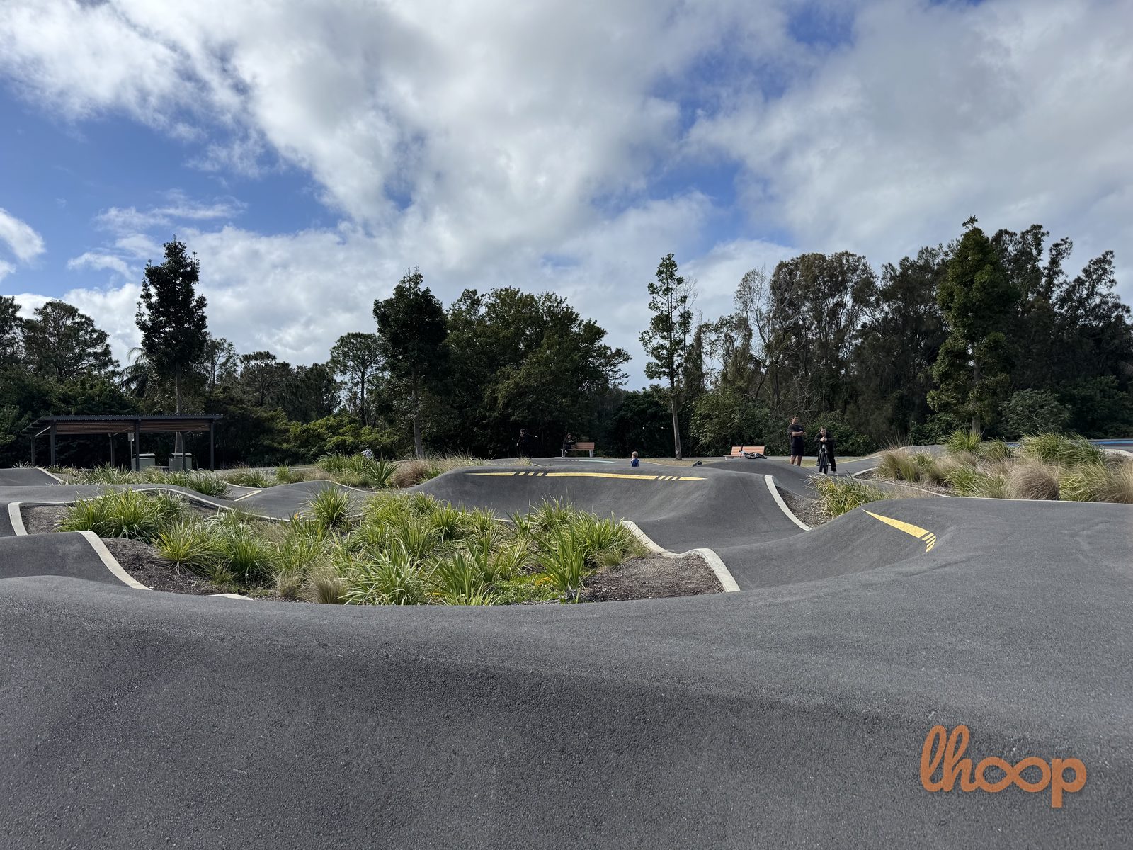 A smooth flowing section of the pump track curving past native plantings, with a shade shelter and riders in the distance.