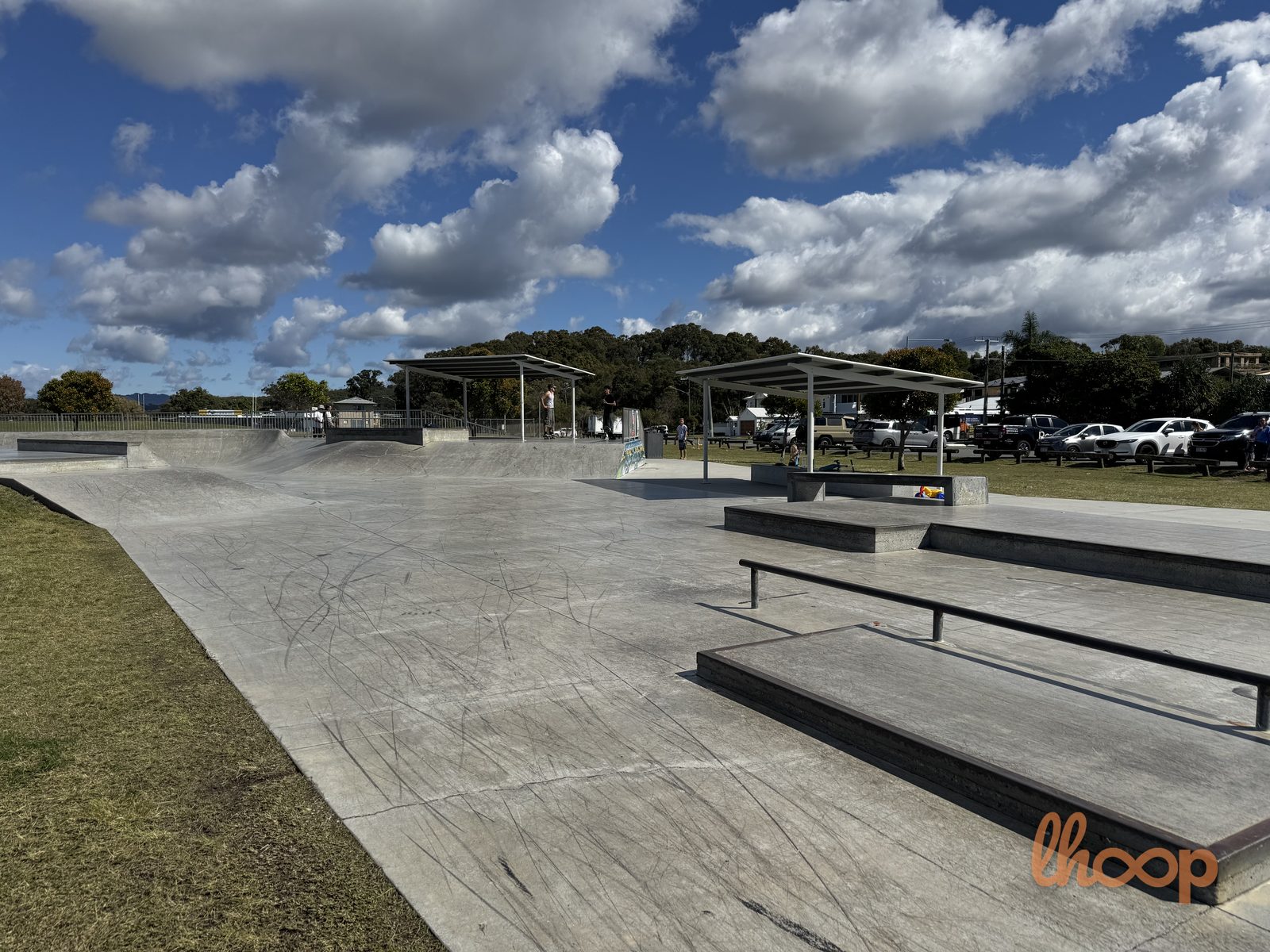 A broad view across the skatepark, with the bowl and older transitions on the left, shade shelters in the middle and the street ledges and stairs on the right.