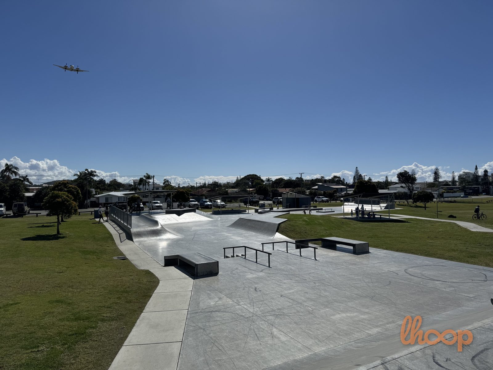The street plaza from above, with flat rails, ledges and manual pads spread across the concrete and a light plane crossing the sky.