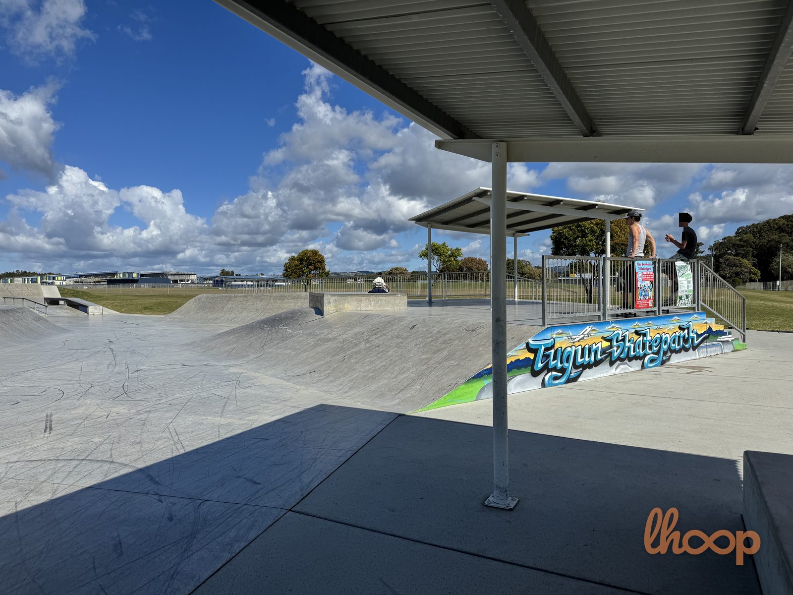 The painted "Tugun Skatepark" mural along a bank, seen from under the viewing shelter with skaters gathered at the rail.