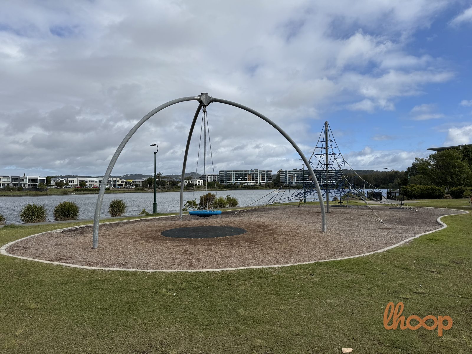 A tall steel arch holding a round nest swing, with a rope climbing pyramid beside it on the Lake Orr foreshore.