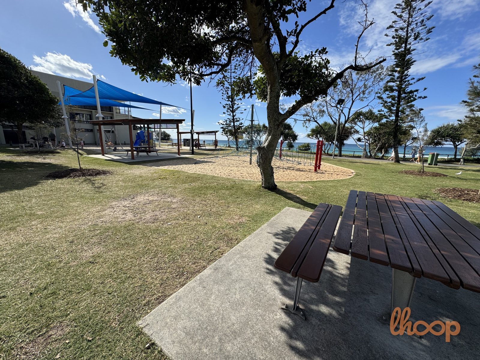The parkland lawn with a shaded picnic table in the foreground and the shade-sail play area and ocean beyond.