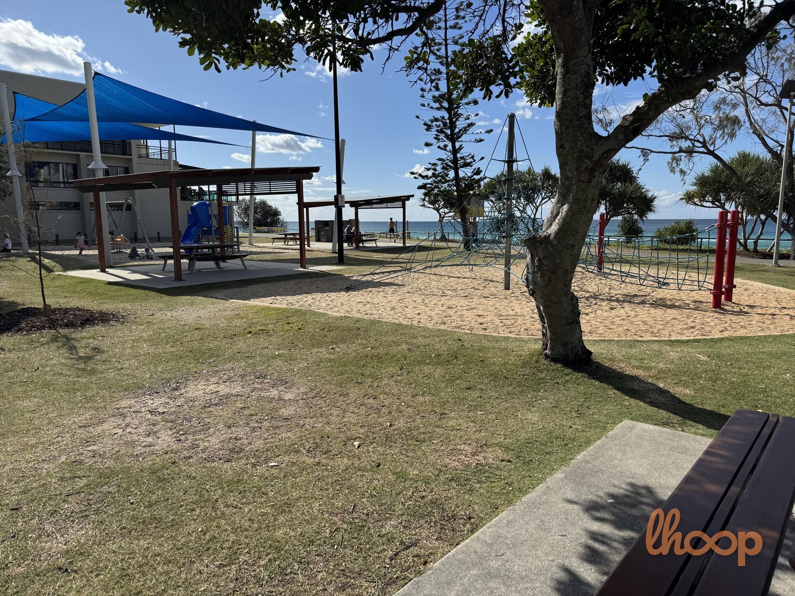 Blue shade sails over a picnic shelter and blue slide unit, with the rope climber and the ocean to the right.