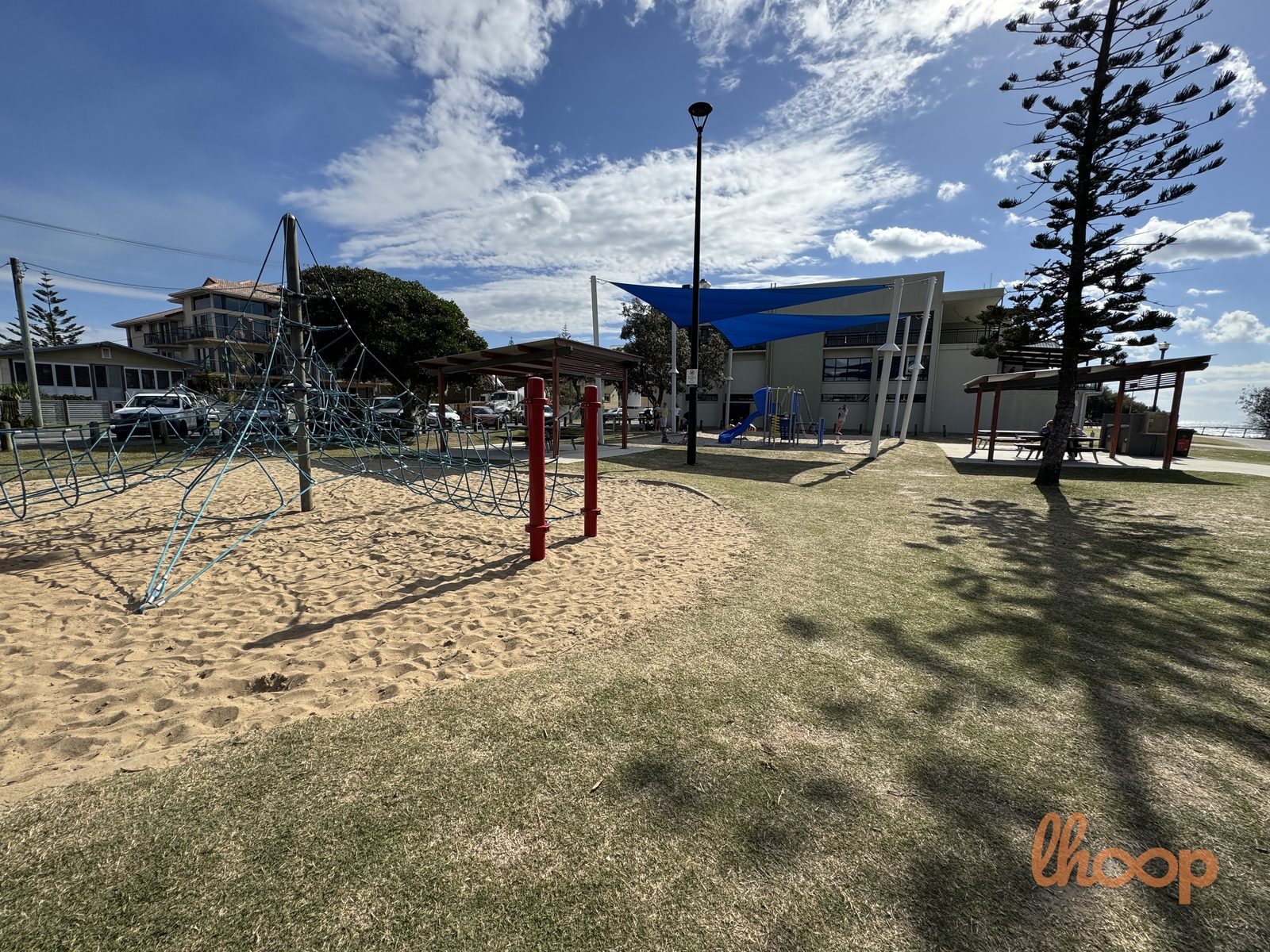 The blue rope pyramid climber on sand, with shade sails, the surf club building and a Norfolk pine behind.